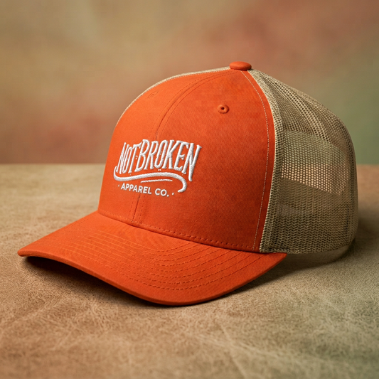 Orange and beige cap with 'Not Broken Apparel Co.' logo on a brown surface